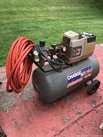 Full view of the air compressor with red air hose coiled on the frame, tank, wheels, and motor housing.