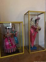 Two glass display cases each containing Asian style dolls; one case with a single pink dressed doll and one case with two dolls in traditional dress.