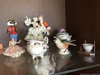 Wide shot of porcelain figurines on a glass shelf including a raccoon, dancing couple, bird lidded bowl, and woman figurine