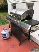 Full view of Char-Broil Classic stainless steel gas grill on wheels with propane tank beside it on red patio.