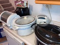 Multiple kitchen appliances together on a kitchen counter including two crock pots (one ceramic with glass lid, one steel with digital control), a can opener, a black grill, an air fryer, and a food chopper.