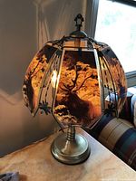 Elk lamp lit up, showing detailed scenic glass panels with elk images surrounding the bulb, metal base, and floral decorative accents on the shade frame.