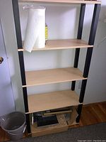 Photo showing particle board shelving unit with 4 shelves, black vertical supports, and light wood-colored shelves. Some miscellaneous items on lower shelves.