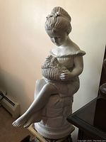 Side view of white statue of girl seated on textured pedestal holding basket