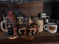 Full view of collection of various German beer steins with some lids, and salt and pepper shakers shaped as steins in a wooden holder.