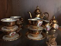 Full view showing complete tea set with teapot, creamer, sugar bowl, cups and saucers, all with gold finish and decorative painting.
