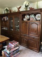Three-piece dark wooden wall unit with multiple storage compartments, some with glass doors and shelving, other with solid wood doors and drawers. Decor items displayed on top and upper shelves including decorative plates and figurines. Stack of board games in front on floor.