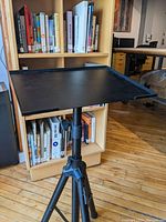 Black adjustable tripod stand with a large rectangular platform on top, placed in front of a bookshelf with books.