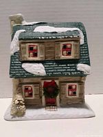 Front view of ceramic Christmas village house showing green roof with snow, red-trimmed windows, wreath on front door, small pine tree on base covered with snow.