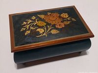 Closed view of the Italian wooden music box with floral inlay on the lid, showing its polished curved body and gold border detail.