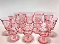 Set of five tall sherbet/champagne pink glasses arranged in back row, and seven water goblets in front, all translucent pink with no chips or cracks.