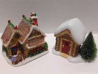 Front angled view of the gingerbread house with candy decoration and the snow-covered log cabin with the tree.