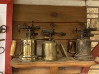 Three antique blow torches arranged on a wooden shelf against a wood-paneled wall.
