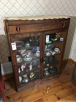 Front view of the vintage wood cabinet with glass doors and drawer containing various small collectibles like ceramic cups, figurines, and glassware.