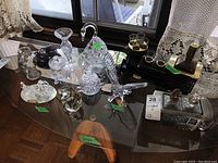 Wide angle photo showing entire lot on glass table with various crystal and glass items plus metal and glass train figurine and decanter sets.