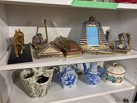 Photo of Egyptian decor on upper shelf including sphinx figurine, desk clock, picture frame, pen stands, and pottery below.