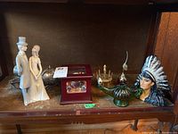 Shelf with assorted decorative items including porcelain bride and groom figurine, red music box, Native American chief head figurine, green glass Aladdin lamp, and brass oyster fork holder.