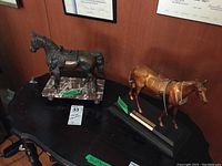 Two metal horse figurines displayed on a dark wood surface; one bronze-colored horse on a black wood base, the other dark metal horse on a stone base.