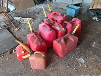 Photo showing nine red plastic jerry cans of varying sizes with yellow pouring spouts attached or black caps. The cans are worn and dirty.
