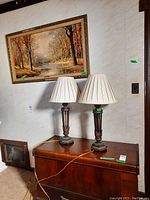Two metal base table lamps with pleated white fabric shades shown on a wooden chest under a painting. Lamps show signs of wear. Shade screws are missing.