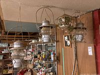 View of three vintage kerosene lanterns hanging from a metal wire frame in a basement workroom with workshop items in background.