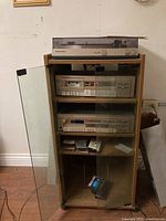 Full system setup in oak-finish cabinet showing turntable on top, cassette deck and tuner behind glass door, tapes on bottom shelf