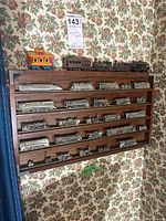 Wall-mounted wooden display shelf with 25 metallic model trains arranged on five rails, plus one colorful wooden caboose on top.