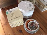 Stack of eight sealed collector plate boxes (five rectangular, three octagonal) alongside a separate stack of unboxed plates.