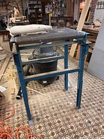 Full view of metal roller stand with canvas tarp folded on top. Blue metal frame with seven horizontally mounted rollers. Basement environment with visible sawdust and wood pieces.