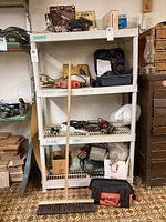 Four shelf plastic shelving unit with various tools including a broom, skill saw, tool bag, hand tools, and other items scattered on shelves in a basement workroom.