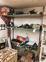 Three metal shelves with various hand tools, power tools, staplers, and tool accessories. Boxes and papers interspersed. Bottom portion shows stacked ceramic tiles and a box of nails on the floor.