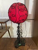 Full view of the vintage spaghetti lamp showing red spherical shade with black wire details on a black metal scrollwork base.