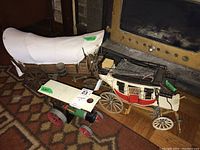 Photo showing three models: a wooden covered wagon with white canvas top, a red and white stagecoach model, and a green and red metal Mamod steam tractor.