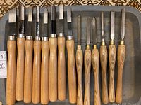 Photo showing seven large and six smaller wood carving chisels arranged in a tray, featuring wooden handles and steel blades in varying widths and shapes.