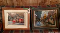 Two framed paintings side by side on a table, showing different scenic artwork in wood frames.