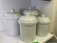Set of four white ceramic canisters with lids placed on a shelf, showing varied heights and clean glossy finish.