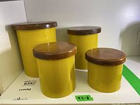 Four yellow Copco metal canisters with wood lids arranged together on a shelf, height range approximately 4.5 to 8 inches tall.