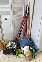 Overview of cleaning supplies lot showing brooms, sponge mop, cleaning liquids, step stool, and other items.