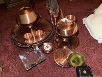 Wide view of copper kitchenware including trays, two pots with lids, small cups, handcrafted MCM plates, small green bowl, and glass ashtray arranged on carpeted floor.