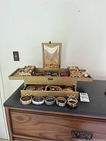 Full view of gold-tone multi-tier jewelry case open with multiple compartments and drawers displaying assorted rings, earrings, bracelets. Several bracelets displayed outside case on tabletop.