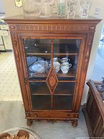Front view of the wooden display cabinet with decorative carved wood and glass door showing inside shelves with dishes.