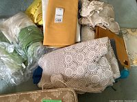 Overview of assorted folded table linens, including crocheted tablecloths, yellow polyester fabric, and wrapped plastic table protectors.