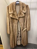 Front view of full-length light brown mink fur coat on hanger, lapel collar, long sleeves and belt.