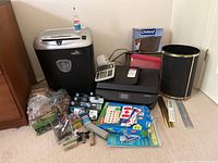 Photo showing Fellowes paper shredder, HP Officejet printer, black waste bin, assorted ink cartridges, pens, folders, and paper clips on carpeted floor.