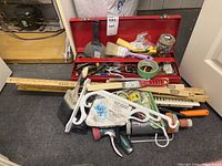 Full view of toolbox opened with hand tools, paint brushes, rolls of tape, yard sticks, and plant hanger visible on floor.