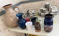 Table with collection of vases including large West German jug-style vase with leaf motif, blue and brown ceramic vase, smaller glass and ceramic vases, and white ceramic vase.
