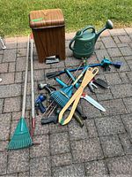Overall lot showing yard tools, trash bin, watering can, garden frog art, hose attachments on patio in sunlight.