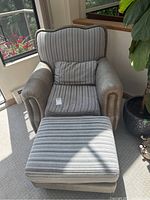 Front view of upholstered armchair with striped cushions and matching ottoman in natural light beside a window and plant.