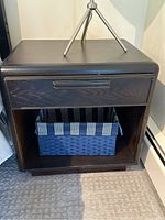 Front view of one dark stained wooden end table showing drawer, lower open shelf with blue striped basket, and visible wear marks.