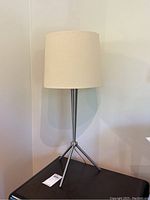 Photo of one silver metal tripod stand table lamp with cream fabric cylindrical shade, shown against a light wall.